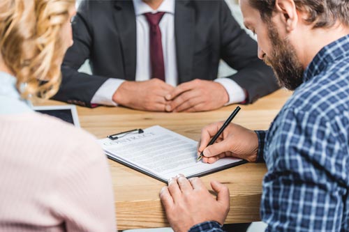 Two  credit union members sit on one side of desk signing a loan document. A bank representative with a suit and tie sits across from them.